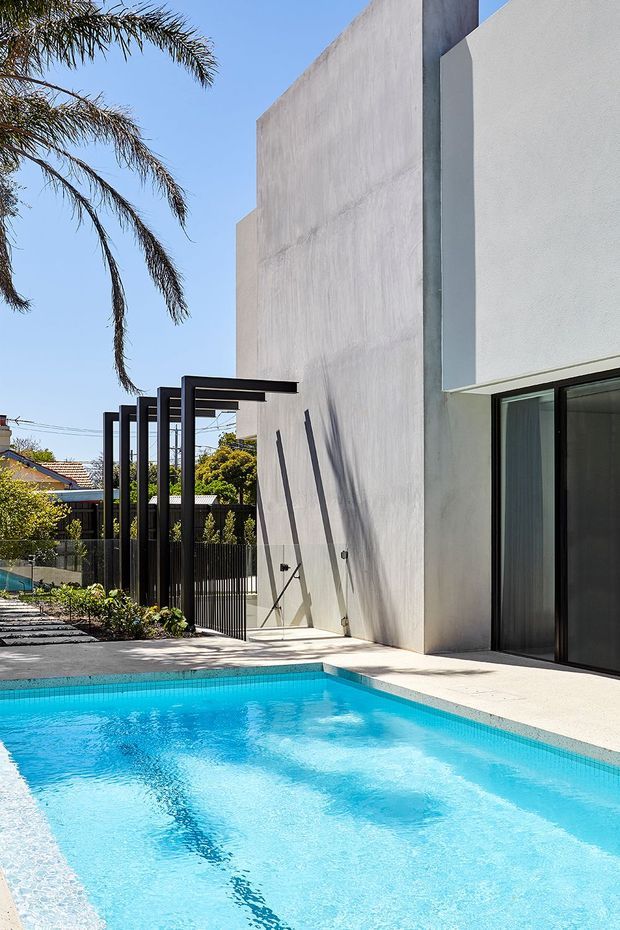 A bright contemporary residential exterior centered on a turquoise swimming pool beside a minimalist white and pale-grey rendered facade. Black-framed floor-to-ceiling glazing opens onto a light concrete terrace, while a sequence of dark steel pergola frames creates a strong linear architectural feature along the side garden. Palm foliage and neighbouring residential rooftops are visible beyond the property.