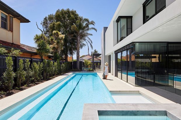 Bright exterior view of a contemporary multi-storey residence with a long rectangular swimming pool along the side of the house. The white rendered building has extensive black-framed glazing and sliding glass doors opening toward the pool, while neighbouring homes and established tropical landscaping border the site.
