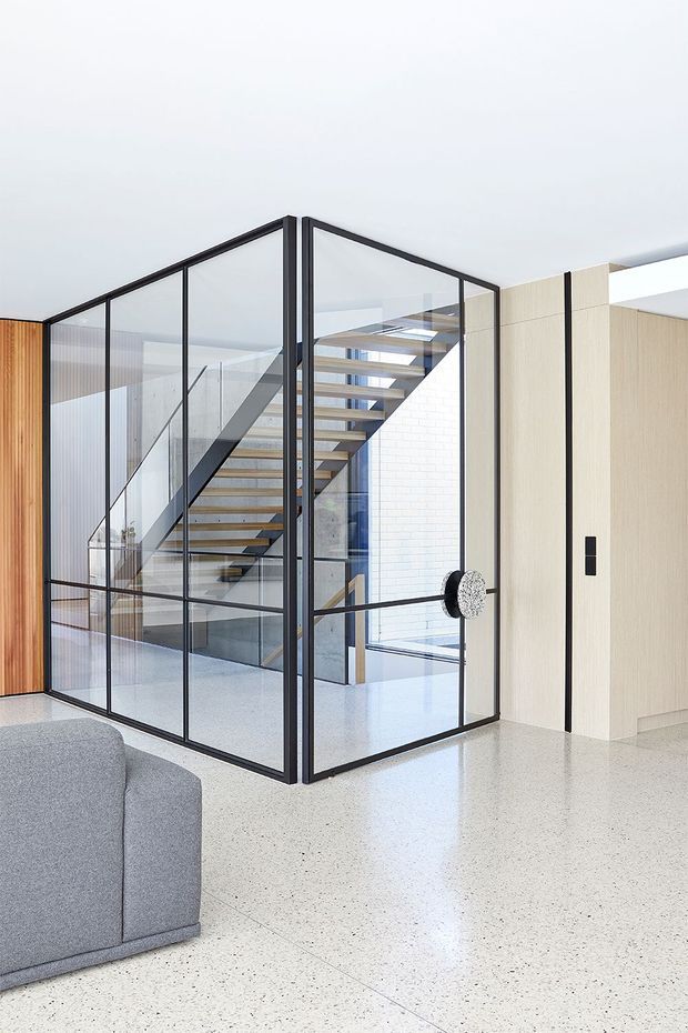 Bright contemporary interior featuring a black-framed glazed stair enclosure with a floating timber staircase. The space has polished light terrazzo flooring, pale timber wall panelling and cabinetry, a white brick wall, and a grey upholstered sofa in the foreground. A sculptural round door handle and open stair void add architectural interest.
