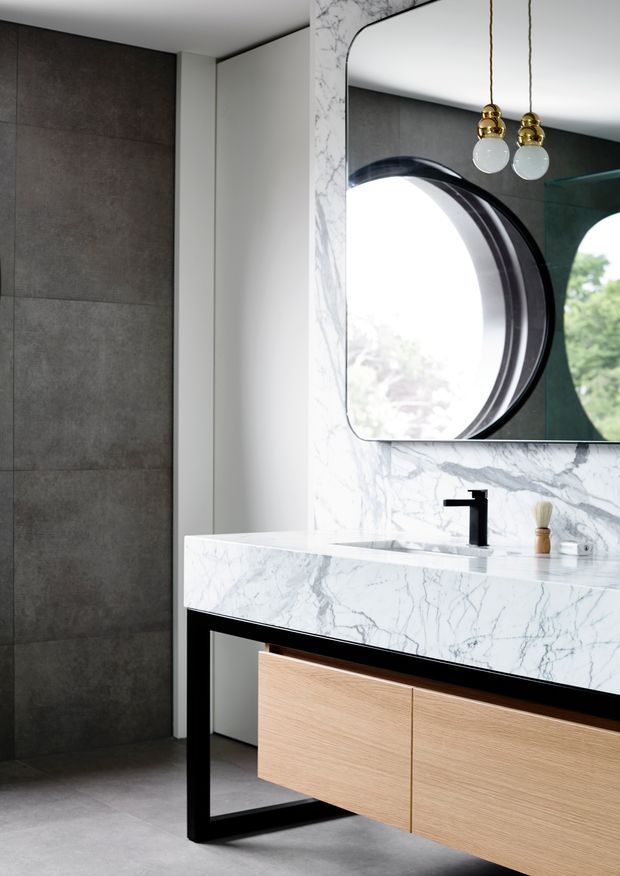 Contemporary bathroom vanity with a floating light-wood drawer unit, thick white marble benchtop and integrated splashback, matte black open-frame supports, black wall-mounted tapware, large rounded-corner mirror, and decorative brass-and-glass pendant lights. Dark large-format wall tiles and grey flooring create a refined monochromatic setting.