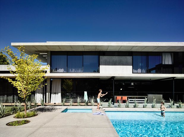 A modern two-storey residence with a strong horizontal concrete roof slab, extensive dark-framed glazing, and a recessed ground-floor outdoor living area facing a swimming pool. The pool terrace includes minimalist landscaping with sculptural grasses and a small tree, while the upper level features large windows and a clean rectangular facade under a clear blue sky.