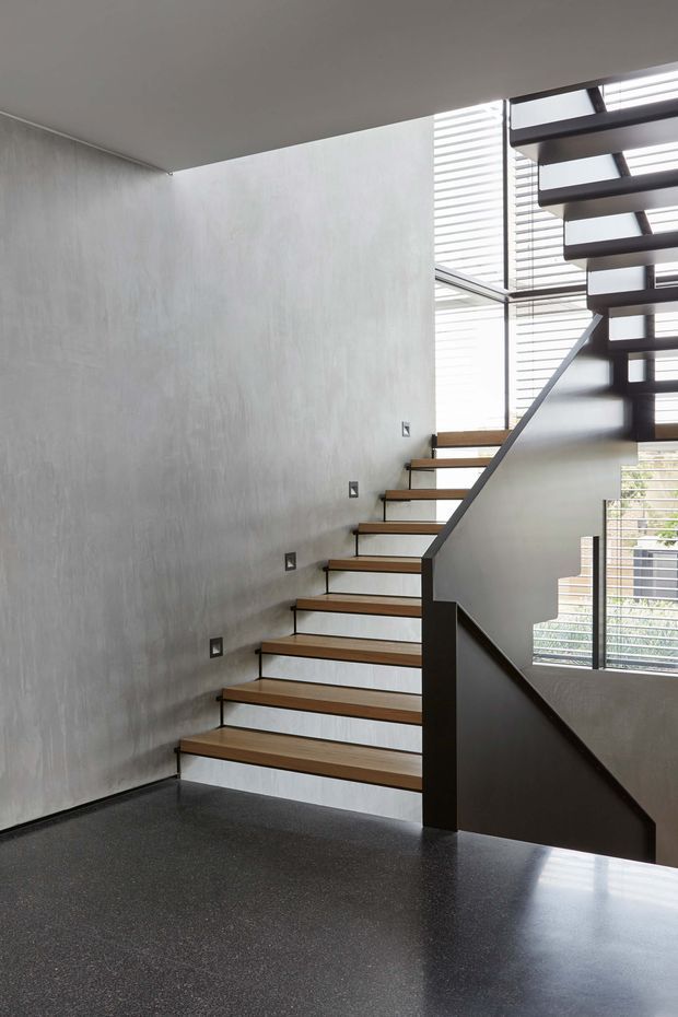 Interior view of a modern open-riser staircase with warm timber treads, dark metal stringers and balustrade, and a grey textured wall. Large glazed openings with horizontal external screens bring in abundant natural light. The lower floor appears dark and polished, with the stair continuing upward out of frame.
