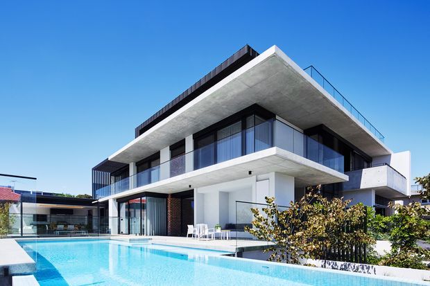A contemporary multi-level residential house viewed from the pool area, featuring strong horizontal concrete slabs, extensive floor-to-ceiling glazing, dark framed windows, open balconies, and a flat roof. A bright swimming pool occupies the foreground, with neighboring low-rise homes visible in the background.