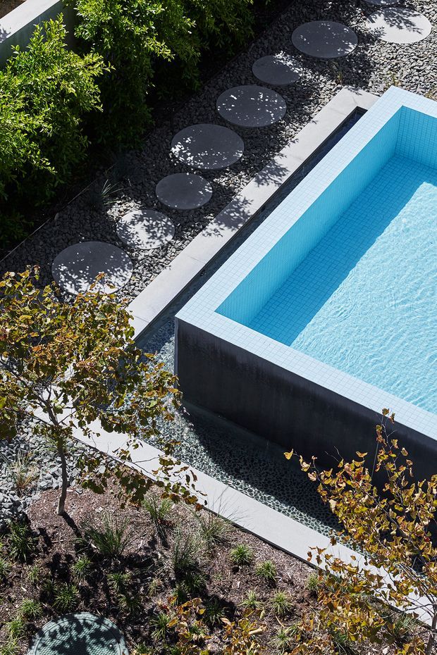 Elevated view of a landscaped outdoor pool area featuring a rectangular swimming pool with bright blue tiled interior, dark exterior walls, and pale tiled coping. A gravel garden path with large circular stepping stones runs alongside dense planting and ornamental trees.