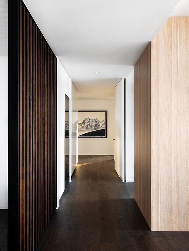 A narrow, refined residential hallway viewed toward an open doorway, with dark timber flooring, a tall dark vertical timber-slat screen on the left, and light timber built-in wall cabinetry or panelling on the right. White walls and ceiling create a bright minimalist backdrop, while a framed monochrome artwork is visible at the end of the corridor.