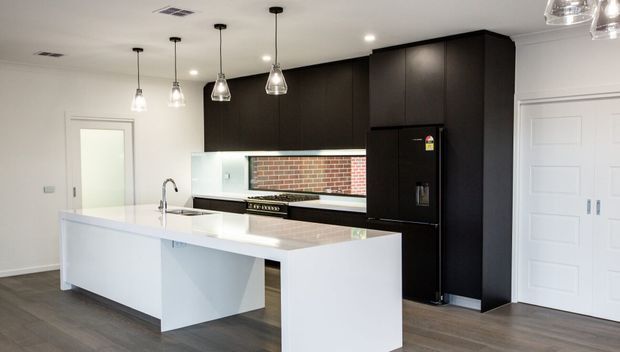 A contemporary kitchen with a large white island and integrated seating overhang, dark matte cabinetry, white benchtops, stainless-steel sink and tapware, and a black refrigerator. A exposed red brick splashback forms a horizontal feature behind the cooktop, while pendant lights hang above the island. The room has light walls, timber-look flooring, and white panelled doors.