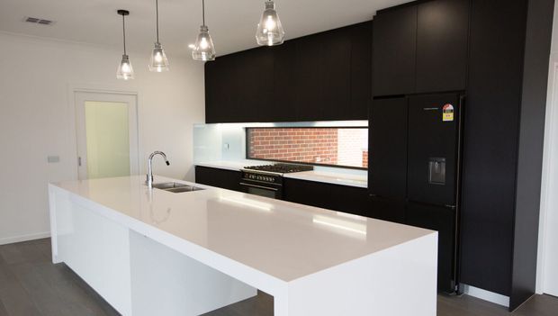 A contemporary kitchen featuring a large white waterfall island with integrated sink and curved faucet, glossy white benchtop, dark full-height cabinetry, a black refrigerator, pendant glass lighting, and a horizontal splashback window revealing exposed red brick outside.