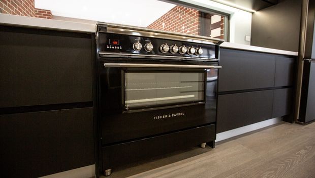 Close-up view of a contemporary kitchen centered on a black freestanding range cooker, set between dark built-in cabinetry. The kitchen features a light wood-look floor, pale benchtop, exposed red brick visible through an opening or glazing, and dark ceiling and wall finishes.