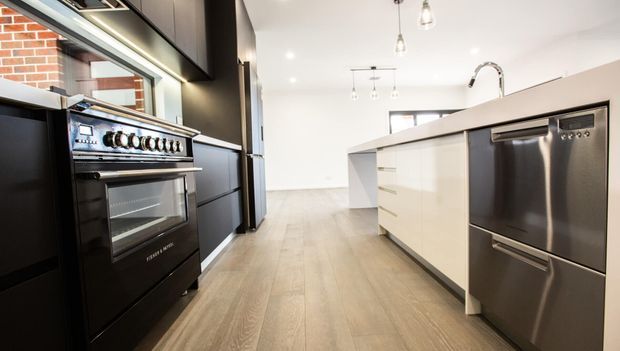 Modern kitchen viewed along a central island, featuring dark cabinetry, a large stainless-steel range cooker and dishwasher, glossy white island cabinetry, light timber-look flooring, pendant lighting, and an exposed brick splashback or wall detail.