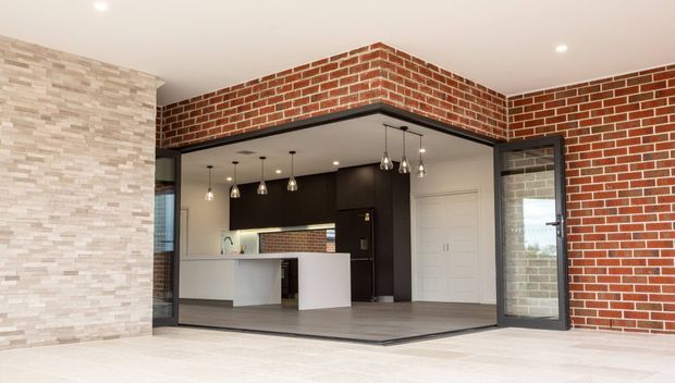 Contemporary open-plan kitchen viewed from a bright adjoining space, framed by exposed red brick walls and a large black-framed glazed opening. The kitchen features dark timber cabinetry, a white island with waterfall-style ends, pendant lighting, pale stone-look wall cladding, and dark flooring.