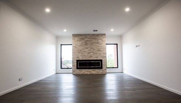 An empty, bright contemporary living room with dark timber-look flooring, white walls and ceiling, recessed downlights, and a central stacked-stone fireplace feature wall flanked by large windows.