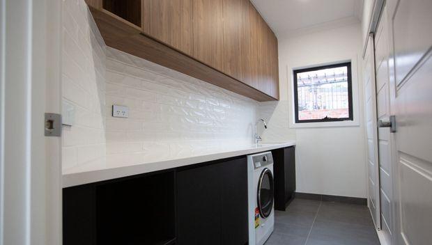 Compact contemporary laundry room with black lower cabinetry, warm wood overhead cupboards, a white textured tiled splashback, pale benchtop, front-loading washing machine, grey floor tiles, and white panelled doors.