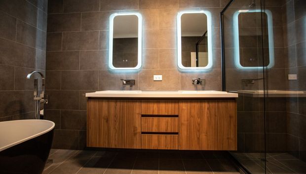 Contemporary dark-tiled bathroom featuring a long timber vanity with two integrated basins, illuminated rounded-rectangle mirrors, a freestanding bathtub, and a frameless glass shower enclosure. The space has a warm, refined, moody atmosphere with strong wood-and-charcoal contrast.