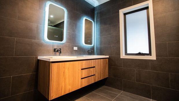 Modern bathroom vanity featuring a floating timber-look double basin cabinet with black tapware, white benchtop, illuminated rounded-rectangle mirrors, and dark large-format wall and floor tiles. A frosted window with a light trim provides natural light.