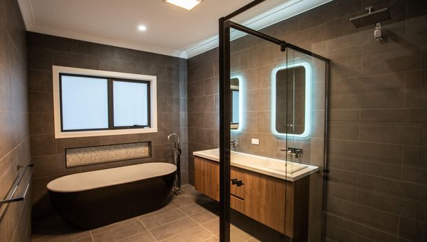 Contemporary bathroom featuring a freestanding oval bathtub, wall-mounted timber vanity with double illuminated mirrors, a glass-enclosed shower, and large dark grey tiled walls and floor. The room has restrained modern detailing with warm wood cabinetry and integrated lighting.
