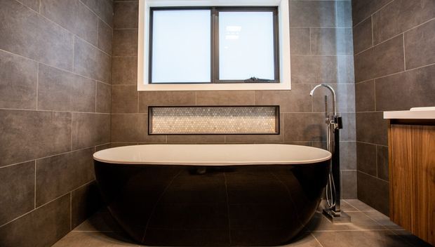 Modern bathroom featuring a freestanding oval bathtub with a dark exterior, large-format dark stone-look wall and floor tiles, a recessed illuminated mosaic niche, a black-framed window, and a timber vanity with a white benchtop. A freestanding chrome bath filler and hand shower sit beside the tub.