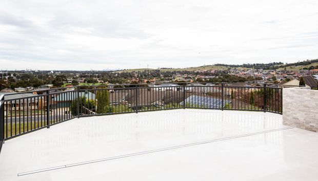 A spacious elevated outdoor terrace or balcony with a glossy light-coloured paved surface, black metal balustrade, and a pale rendered masonry feature wall. The terrace overlooks a suburban residential landscape with rolling hills under an overcast sky.
