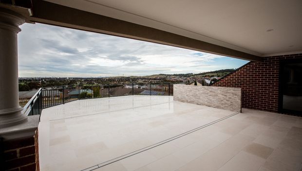 A spacious covered outdoor terrace with pale tiled flooring, black metal balustrades, a classical column, and a partial red-brick wall. The elevated terrace overlooks a suburban landscape and rolling hills beneath a cloudy sky.