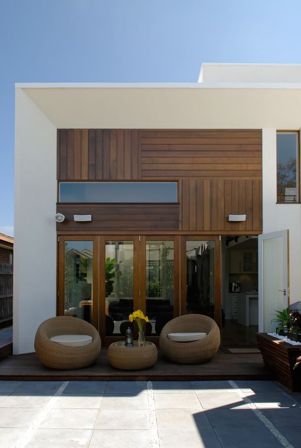 A contemporary two-storey residential facade opening onto a paved courtyard or terrace. The white rendered exterior has strong horizontal rooflines and a projecting upper volume clad in dark timber boards. Timber-framed glazed doors connect the interior to the outdoor seating area, with two rounded woven lounge chairs and a matching low table on a dark timber deck. An open side door reveals part of the interior, while large pale paving slabs occupy the foreground.