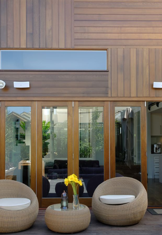 A warmly finished indoor seating area facing a wall of timber-framed glazed doors and windows. Vertical and horizontal timber wall panelling surrounds a high clerestory-style window, while two rounded woven lounge chairs and a matching low circular table sit on dark timber flooring. Through the glazing and reflections, a darker sofa, greenery, and part of a kitchen are visible. The space has abundant natural light and a relaxed, resort-like atmosphere.