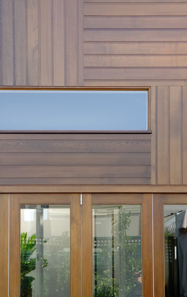 Close-up exterior architectural view of a contemporary timber-clad facade. The wall combines vertical and horizontal timber boards with a narrow horizontal window and glazed timber-framed doors below. Reflections and glimpses of greenery are visible through the glazing, but the main subject is the building envelope and its detailed timber composition.