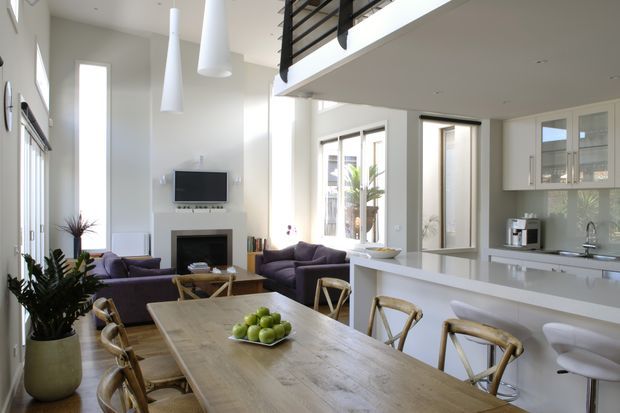 Bright open-plan interior combining a dining area, living room, and kitchen. A large timber dining table with woven-back chairs occupies the foreground, while a white kitchen with peninsula seating is visible to the right. The living area includes purple sofas, a fireplace, and wall-mounted television. Tall windows and doors bring in abundant daylight, and a mezzanine or upper-level opening with dark metal balustrading is visible above.