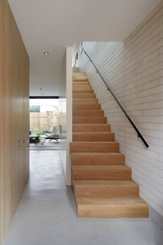 A bright contemporary interior hallway centred on a straight timber staircase. The stair has broad light-oak treads and risers, a slim black metal handrail, and a white painted brick wall along one side. Pale concrete flooring, vertical timber wall panelling, and a glazed opening toward a softly furnished living area create a restrained, warm minimalist composition.