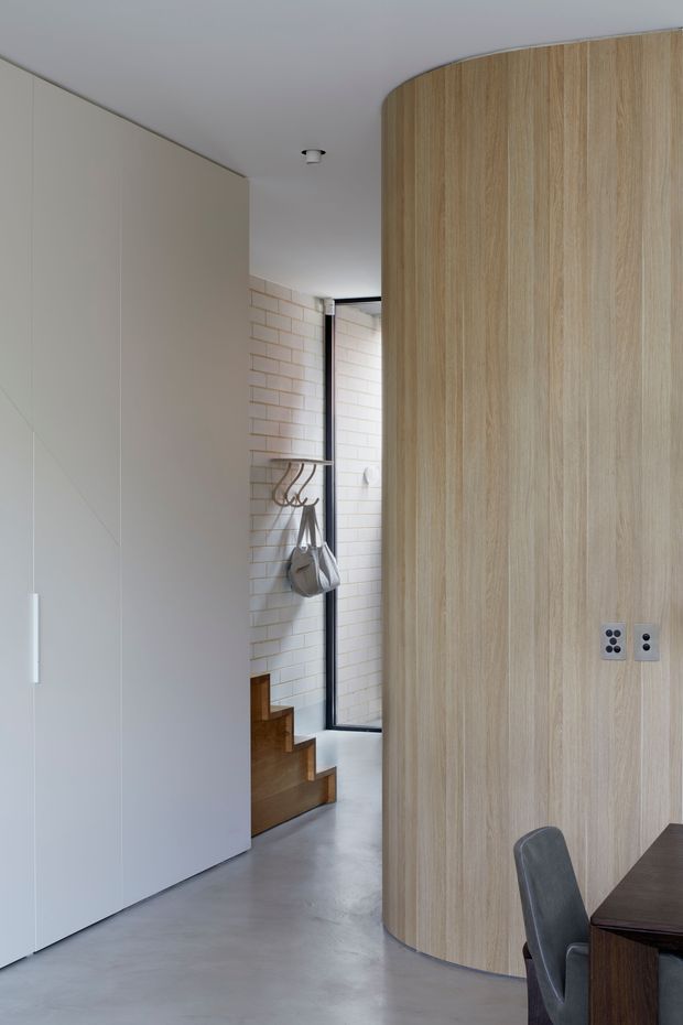 A refined contemporary interior view of a circulation and dining area. A curved, floor-to-ceiling light timber veneer partition or built-in wall dominates the right side, with integrated electrical outlets and a dark dining table partly visible. A large flush white built-in cabinet lines the left wall. Beyond the partition is a tiled entry or stair zone with sculptural hooks, a small timber stair, black-framed glazing, and a polished grey concrete floor. The space uses clean lines, concealed joinery, and a restrained neutral palette.