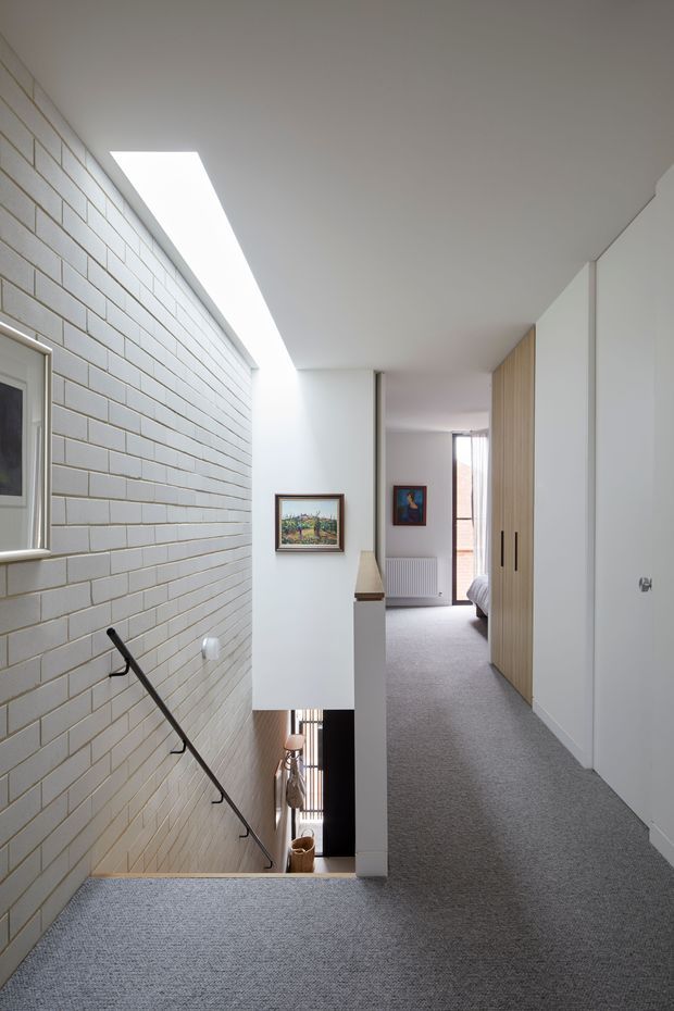A bright, minimalist upper-floor hallway and stair landing. The space features grey carpet flooring, a white-painted ceiling and walls, a long skylight, a pale glazed brick stairwell wall with a slim black handrail, and built-in light timber and white wardrobes. The corridor leads toward a partially visible bedroom, with framed artwork and a radiator adding detail.
