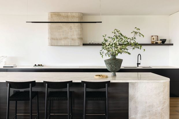A refined contemporary kitchen viewed straight on, featuring a long dark island with three black stools, a pale stone waterfall end, a dark benchtop, white cabinetry or wall surfaces, and a minimalist black linear pendant. A sculptural ribbed cylindrical rangehood or hood enclosure hangs above the cooking area, while open shelving displays restrained decor and a vase of foliage provides a natural focal point.