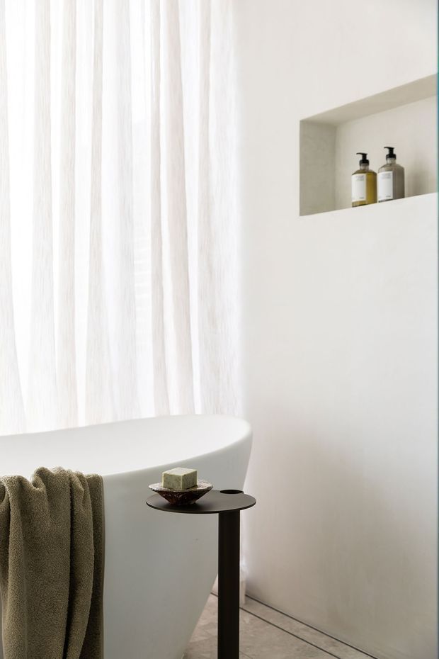 A serene, minimalist bathroom featuring a freestanding white bathtub beside sheer white curtains. A recessed wall niche holds two dark pump bottles, while a slim black side table displays soap and a small dish. The space has pale walls, a light stone or concrete-look floor, and soft natural light.