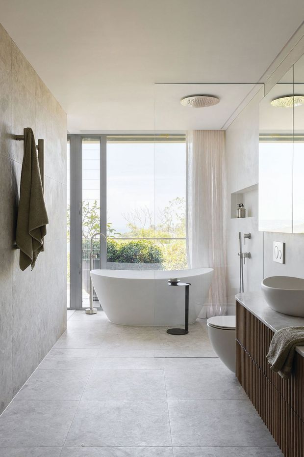 Bright contemporary bathroom with a freestanding white bathtub positioned in front of full-height glazing, a walk-in shower area, wall-mounted toilet, round vessel basin, large mirror, and vertically ribbed timber vanity cabinetry. The room features pale large-format floor and wall tiles, soft sheer curtains, integrated ceiling shower fittings, and abundant natural light overlooking planted greenery.