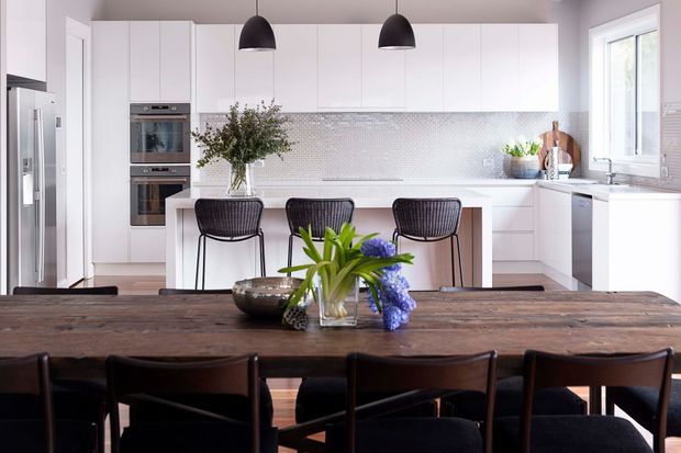 Bright contemporary kitchen and dining area with white flat-panel cabinetry, integrated stainless-steel appliances, a long island with three dark woven bar stools, and a large rustic timber dining table in the foreground. Black pendant lights and a pale geometric tile splashback provide contrast, while a large window brings in natural light.