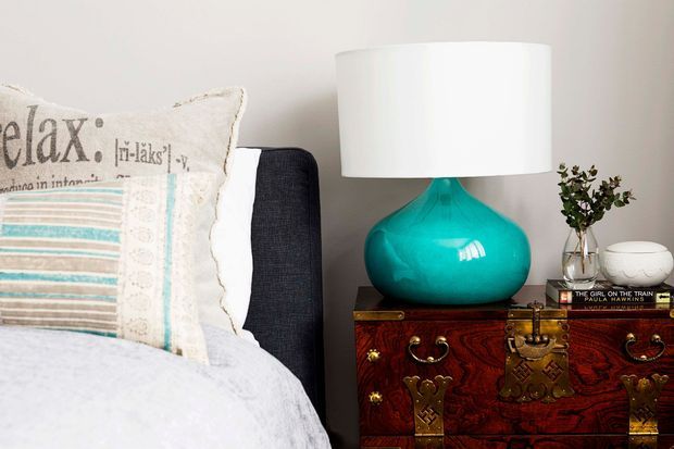 Close-up bedroom vignette featuring a dark upholstered bed with layered decorative pillows and pale bedding beside a richly carved dark wood chest used as a bedside table. A vivid turquoise table lamp, white drum shade, small vase of foliage, and decorative objects add colour and character against a light neutral wall.