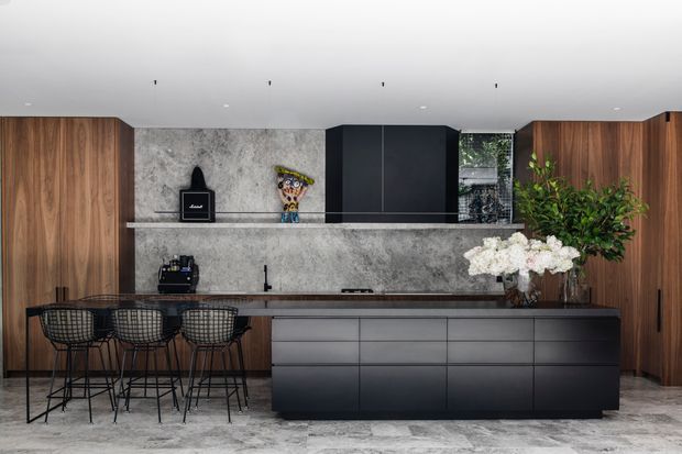 A sophisticated contemporary kitchen with a long central island, dark charcoal cabinetry, warm vertical timber wall cabinetry, and a grey veined stone backsplash and floor. Three black wire bar stools line the island, while integrated appliances and open shelving are arranged along the back wall. Minimal recessed ceiling lighting, indoor greenery, and floral arrangements add a refined, modern feel.