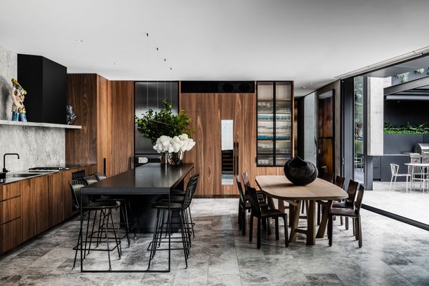 A spacious contemporary open-plan kitchen and dining area with dark timber cabinetry, a black stone island, integrated appliances, and a large dining table. Concrete-look flooring and walls create a refined industrial feel, while full-height glazing opens the room to an outdoor courtyard or terrace. Black-framed glass doors, pendant lighting, and built-in display shelving complete the interior.