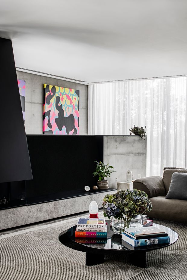 A refined contemporary living room with a large black built-in media unit against a raw concrete wall, floor-to-ceiling sheer curtains, a low round black coffee table, and a taupe sectional sofa. Colourful abstract artwork, indoor plants, books, flowers, and sculptural accessories add contrast to the monochrome concrete and dark joinery. The room has a bright, soft-lit atmosphere with strong geometric built-in elements and a layered, curated interior.