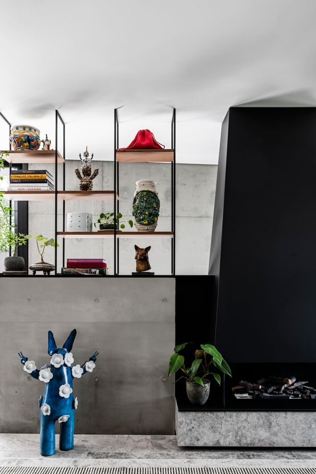 A contemporary living room featuring a dramatic black geometric fireplace wall and hearth, a raw concrete partition, and slim black-framed open shelving displaying books, ceramics, and sculptural objects. A vivid blue decorative sculpture, potted plants, and a textured pale stone or concrete floor add contrast to the monochrome interior.