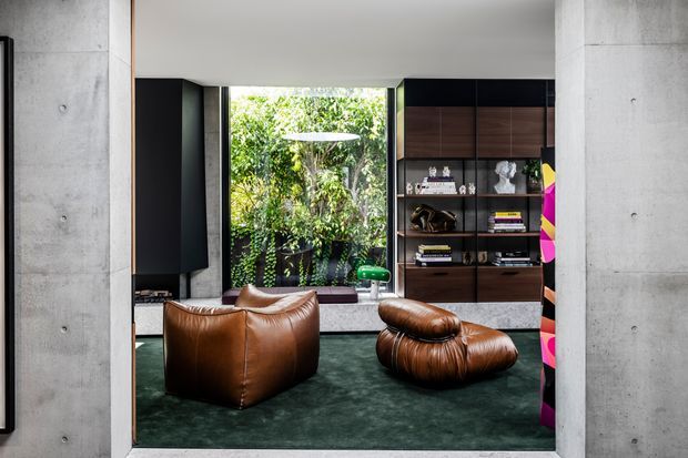 A sophisticated modern living room framed by raw concrete walls, with rich brown leather lounge chairs and ottoman seating on a deep green carpet. A full-height glazed opening looks onto dense greenery, while dark built-in shelving and warm timber cabinetry display books and objects. The space has a refined, moody, gallery-like atmosphere with strong indoor–outdoor connection.