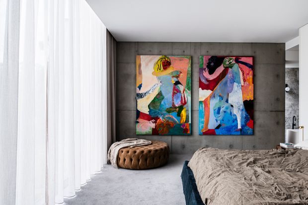 A contemporary bedroom with floor-to-ceiling sheer curtains, grey carpet, a dark concrete-look feature wall, and two colourful abstract artworks above a tufted round ottoman. A partially visible bed with textured grey bedding occupies the foreground, while a compact vanity or bathroom area appears at the right edge.