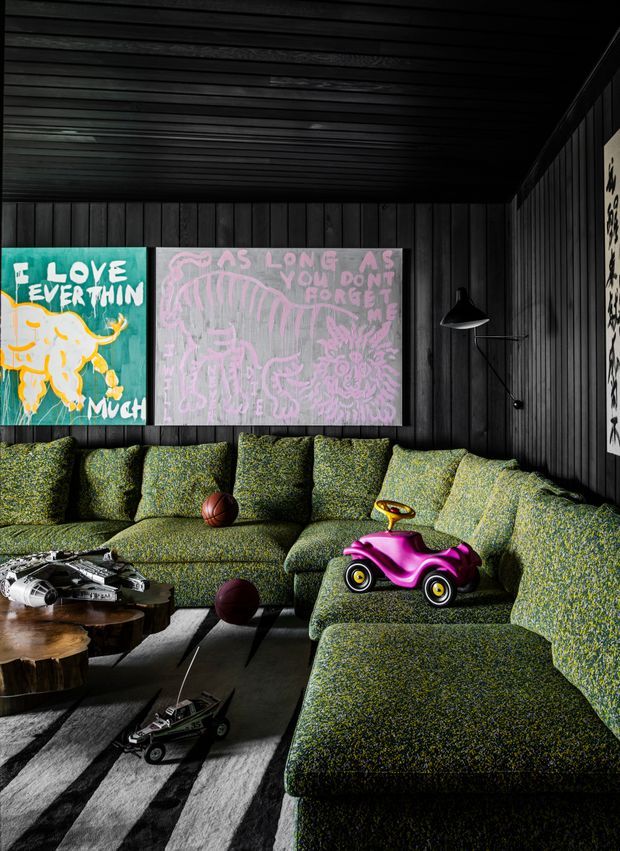 A dark, moody lounge room with black vertical timber wall panelling and a matching dark slatted ceiling. A large textured green sectional sofa wraps around the room, accompanied by a black floor lamp, low organic timber coffee tables, a striped rug, children's toys, and colourful playful artwork mounted on the wall.