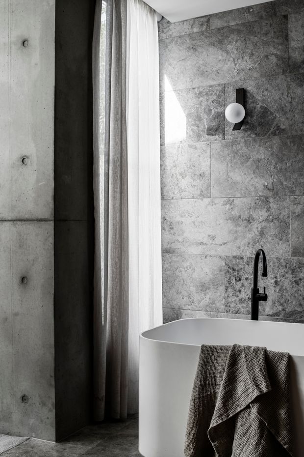 A monochrome contemporary bathroom featuring a freestanding white oval bathtub, black wall-mounted tapware, a black-and-white spherical wall light, large grey stone-look wall tiles, and an exposed concrete wall beside full-height sheer curtains. The space has a restrained, high-contrast minimalist aesthetic with natural light.