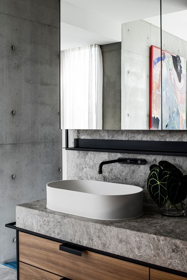 Close-up contemporary bathroom vanity with a white vessel basin set into a thick grey stone benchtop, black wall-mounted tapware and shelf, timber drawer cabinetry, and a large mirror. The space features exposed concrete walls and columns, sheer curtains, and a colourful artwork reflected beside the mirror, creating a stark minimalist industrial aesthetic.