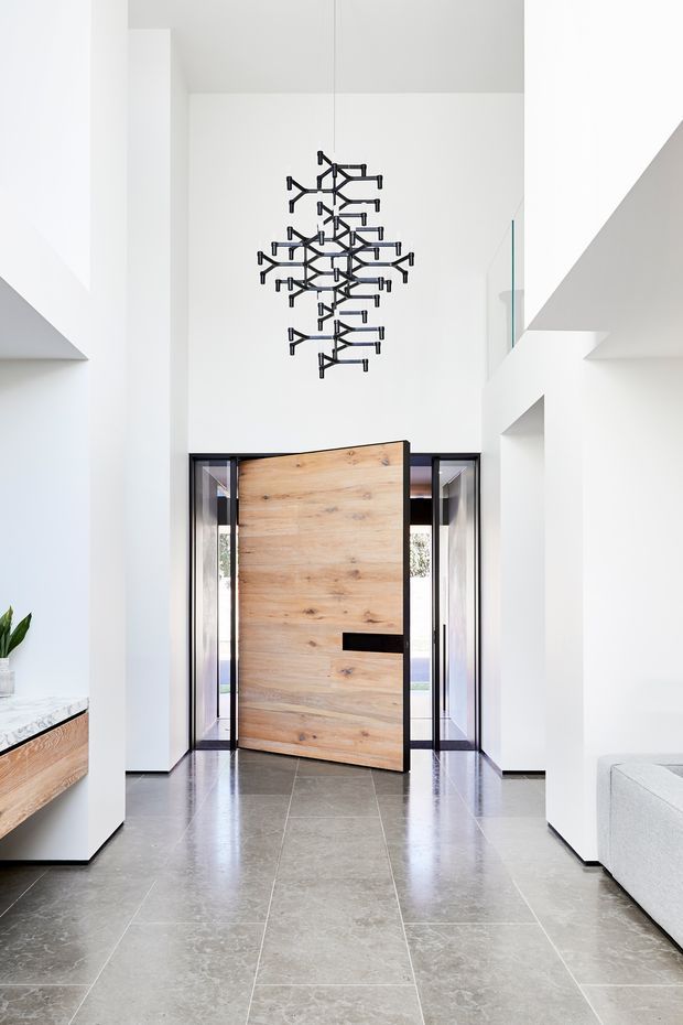Bright, minimalist residential entryway with a dramatic double-height void. A large natural timber pivot door is set within dark-framed glazing, opening to the exterior. Large grey stone-look floor tiles, white walls, angular built-in architectural forms, and a sculptural black branching pendant create a clean contemporary composition. Partial built-in timber joinery and a light upholstered sofa are visible at the sides.