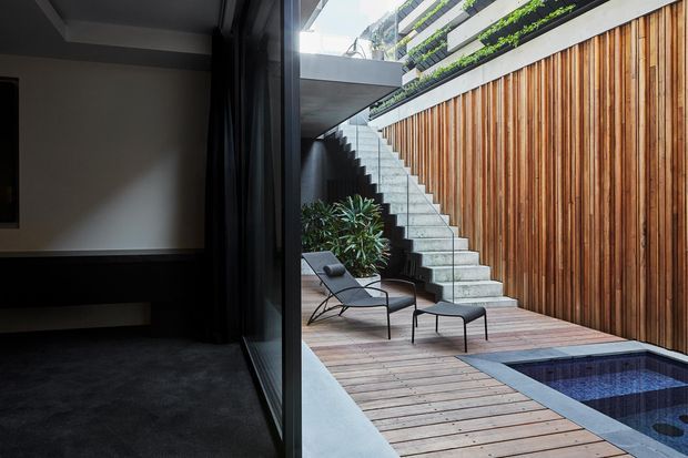 A contemporary urban courtyard viewed from a dark interior through full-height glazing. The courtyard features a timber-lined wall, exposed concrete stairway, timber decking, a compact tiled plunge pool, and minimalist outdoor lounge furniture. Layered green planting is visible on the upper level, creating a private, sculptural outdoor space.