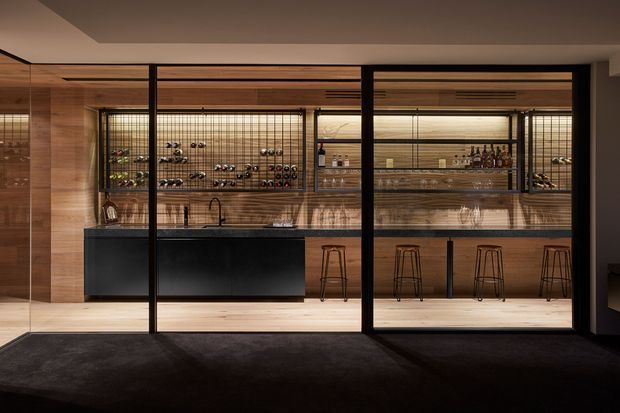 A sophisticated home bar viewed through black-framed glass partitions. The room features warm timber wall and ceiling finishes, dark built-in cabinetry, a stone-look benchtop, open wine storage, integrated shelving with bottles and glassware, and a long bar counter with multiple stools. The low, warm lighting creates a refined and moody atmosphere.
