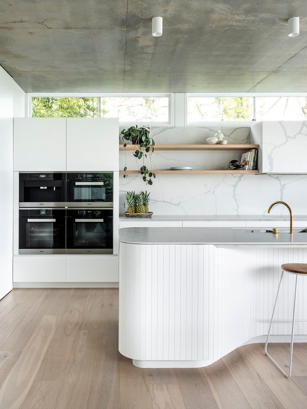 Bright contemporary kitchen featuring a large curved island with vertically fluted white cabinetry, a pale stone benchtop and matching marble-look splashback. White full-height cabinetry houses multiple black built-in ovens, while open timber shelving displays decor and books. Light timber flooring, horizontal clerestory windows, a raw exposed concrete ceiling, and brushed brass tapware complete the clean, refined space.