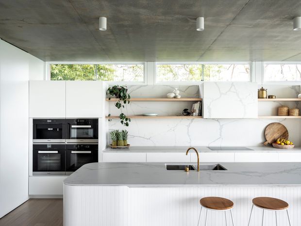 Bright contemporary kitchen with a large white waterfall island featuring vertically fluted detailing, two timber-seated stools, integrated black appliances, open timber shelving, white cabinetry, and a veined marble-look splashback and benchtop. A raw concrete ceiling with simple cylindrical pendant lights contrasts with the light, refined joinery. High clerestory-style windows bring in greenery-filtered daylight.