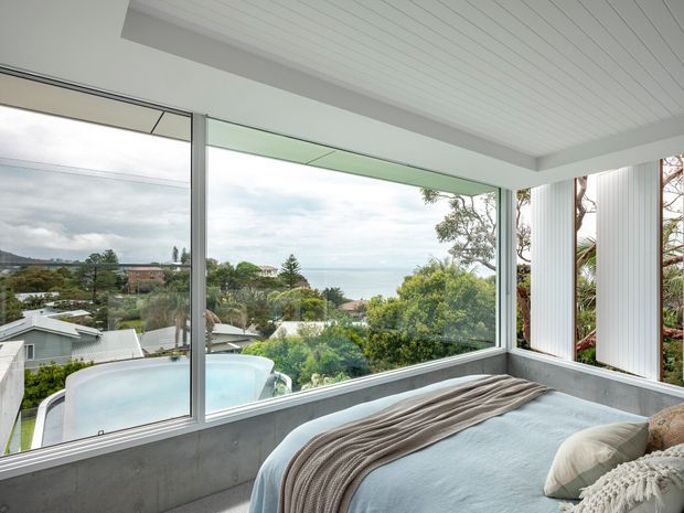 A bright contemporary bedroom corner with a bed positioned beside expansive corner glazing. The room has white vertical wall panelling, a white-lined ceiling, pale bedding, and a grey concrete-look lower wall. The windows overlook a leafy suburban coastal landscape, with a swimming pool visible below.