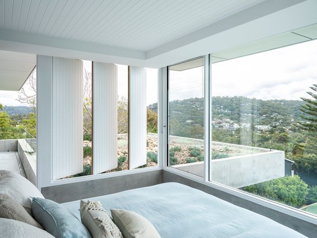 A bright bedroom with a low-profile upholstered bed positioned beside expansive corner glazing. The room features a white ribbed ceiling, pale neutral bedding, exposed concrete window surrounds and planter walls, and vertical white privacy fins. The glazing overlooks a lush, hilly suburban landscape, while a narrow planted terrace is visible outside.